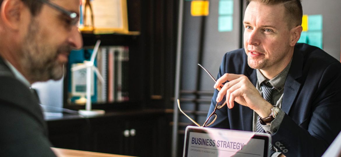 Two men in a modern office discuss business strategy, with colorful sticky notes on the wall and a laptop on the table displaying charts and graphs. One man holds glasses, listening attentively as they uncover content-missing elements crucial to their project.