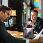 Two men in a modern office discuss business strategy, with colorful sticky notes on the wall and a laptop on the table displaying charts and graphs. One man holds glasses, listening attentively as they uncover content-missing elements crucial to their project.