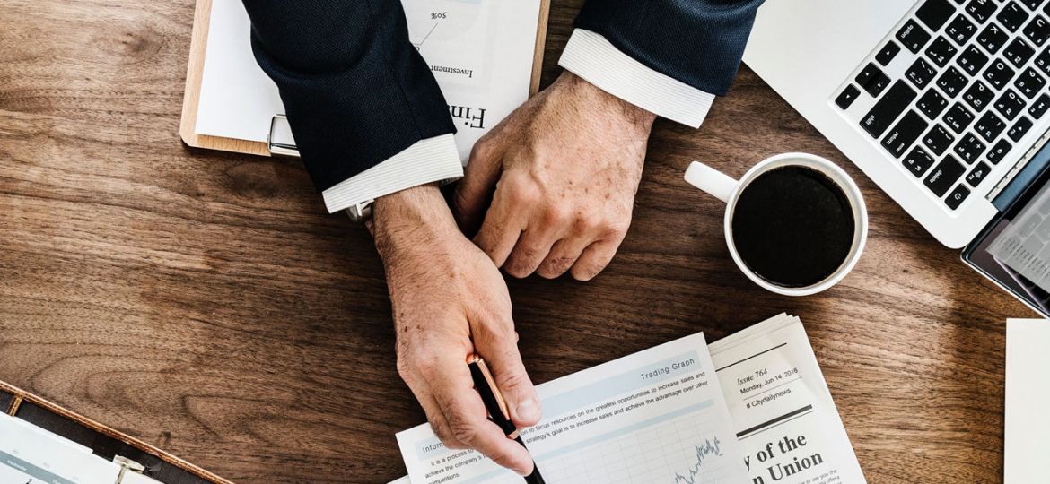 A person in a suit is sitting at a desk covered with business documents and charts, pointing with a pen at a graph on a piece of paper. Despite the wealth of information, the content-missing sections stand out. Nearby are a laptop, cup of coffee, newspapers, and an open planner.