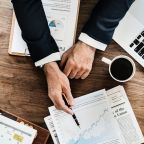 A person in a suit is sitting at a desk covered with business documents and charts, pointing with a pen at a graph on a piece of paper. Despite the wealth of information, the content-missing sections stand out. Nearby are a laptop, cup of coffee, newspapers, and an open planner.