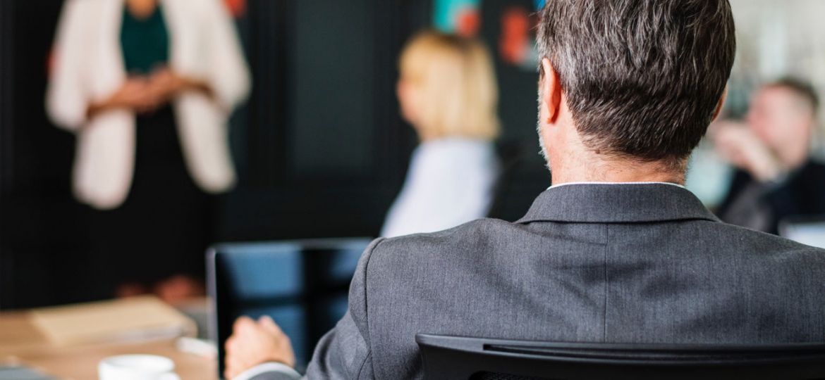 A man in a gray suit sits at a table, facing blurred coworkers during a meeting. A woman stands in front, speaking, with colorful sticky notes on the black wall behind her—capturing essential details to avoid content-missing moments. A cup and tablet are on the table.
