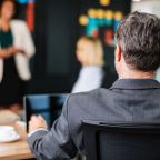A man in a gray suit sits at a table, facing blurred coworkers during a meeting. A woman stands in front, speaking, with colorful sticky notes on the black wall behind her—capturing essential details to avoid content-missing moments. A cup and tablet are on the table.