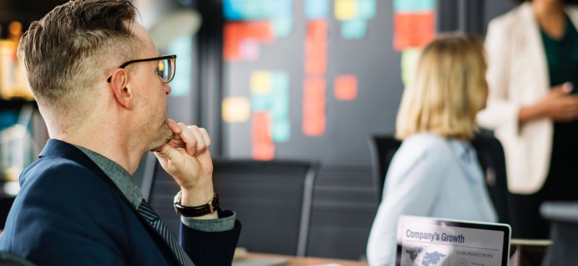 A professional meeting setting with a man in a suit seated in the foreground, looking towards a woman standing near a colorful wall of sticky notes. A laptop displaying a "Company's Growth" presentation is on the table, subtly hinting at content-missing slides yet to be completed.