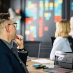 A professional meeting setting with a man in a suit seated in the foreground, looking towards a woman standing near a colorful wall of sticky notes. A laptop displaying a "Company's Growth" presentation is on the table, subtly hinting at content-missing slides yet to be completed.