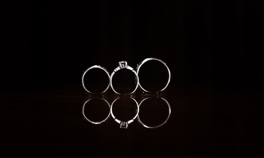 Three rings, including a solitaire engagement ring in the center, stand upright against a black background. Their reflections are visible on the shiny surface below.