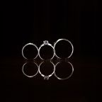 Three rings, including a solitaire engagement ring in the center, stand upright against a black background. Their reflections are visible on the shiny surface below.