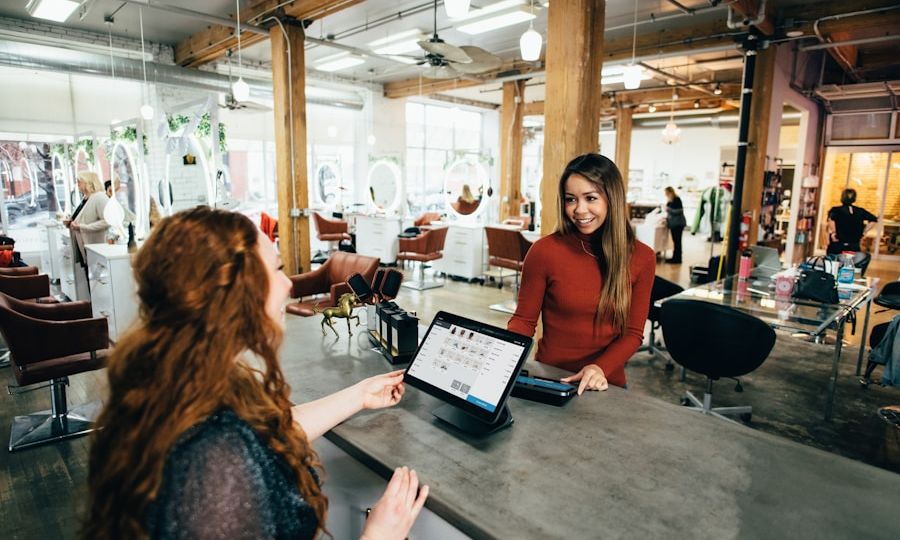 Two women smiling and talking at a salon reception desk. One woman is behind a counter with a computer screen, while the other is facing her, gesturing with her hand. The salon interior is spacious with wooden beams and stylish furniture.