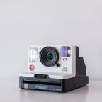 A vintage-style Polaroid OneStep 2 instant camera in white and black sits on a white surface against a plain background. The camera features visible lens, flash, red shutter button, and rainbow detail.
